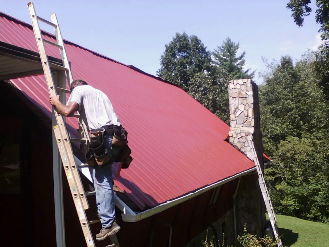 Roofer on a ladder beside a red metal roof and stone chimney — Soffit & Fascia Repair by Roofing Brothers in New Hartford